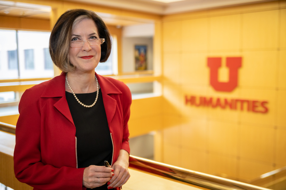 Dean Hollis standing in front of the Humanities block U in the LNCO building. She is wearing a red blazer, black top, and pearl necklace. 