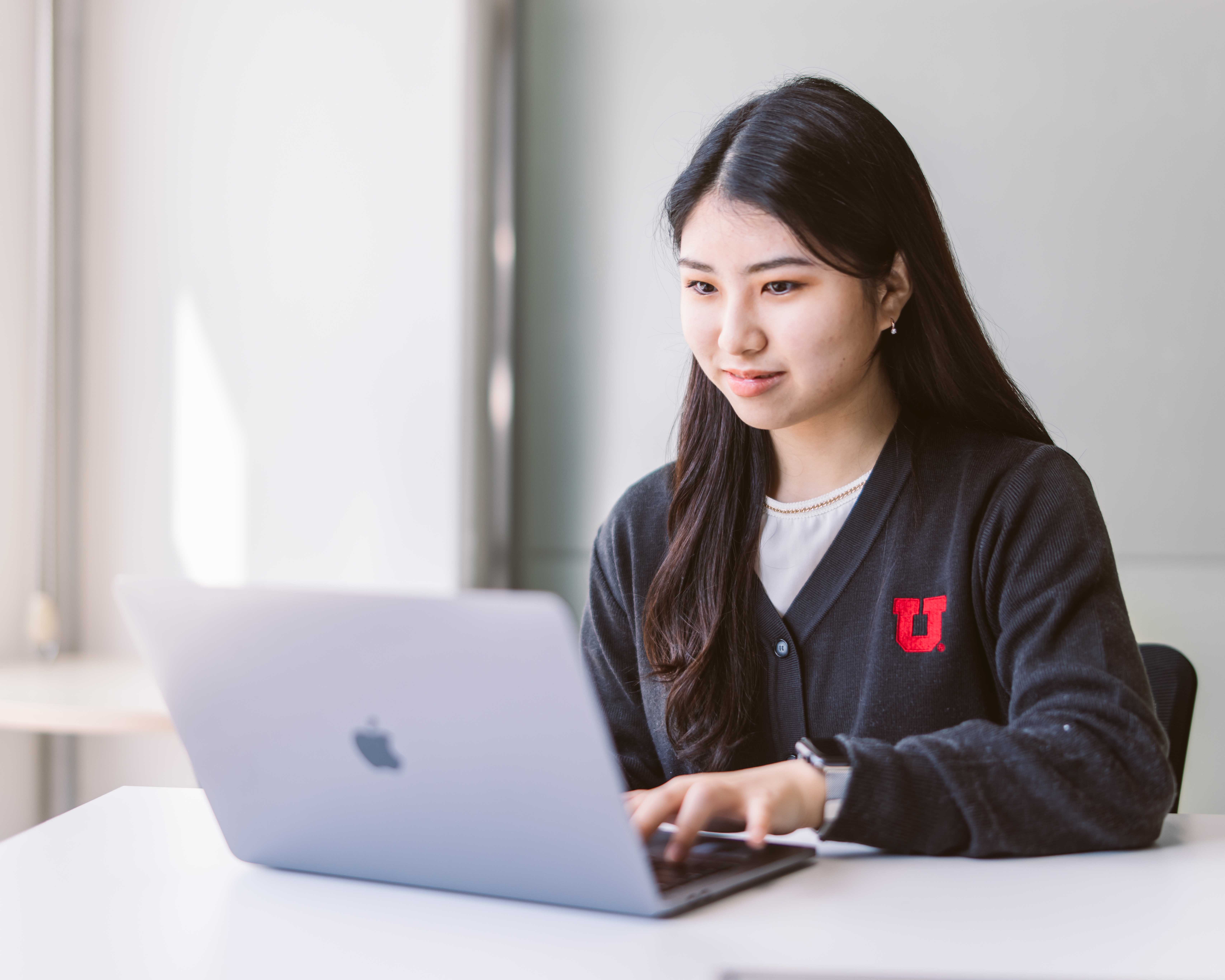 A female student seats at a desk with a silver Mac. She has long dark hair and is wearing a black cardigan with and block U in red on the cardigan. 