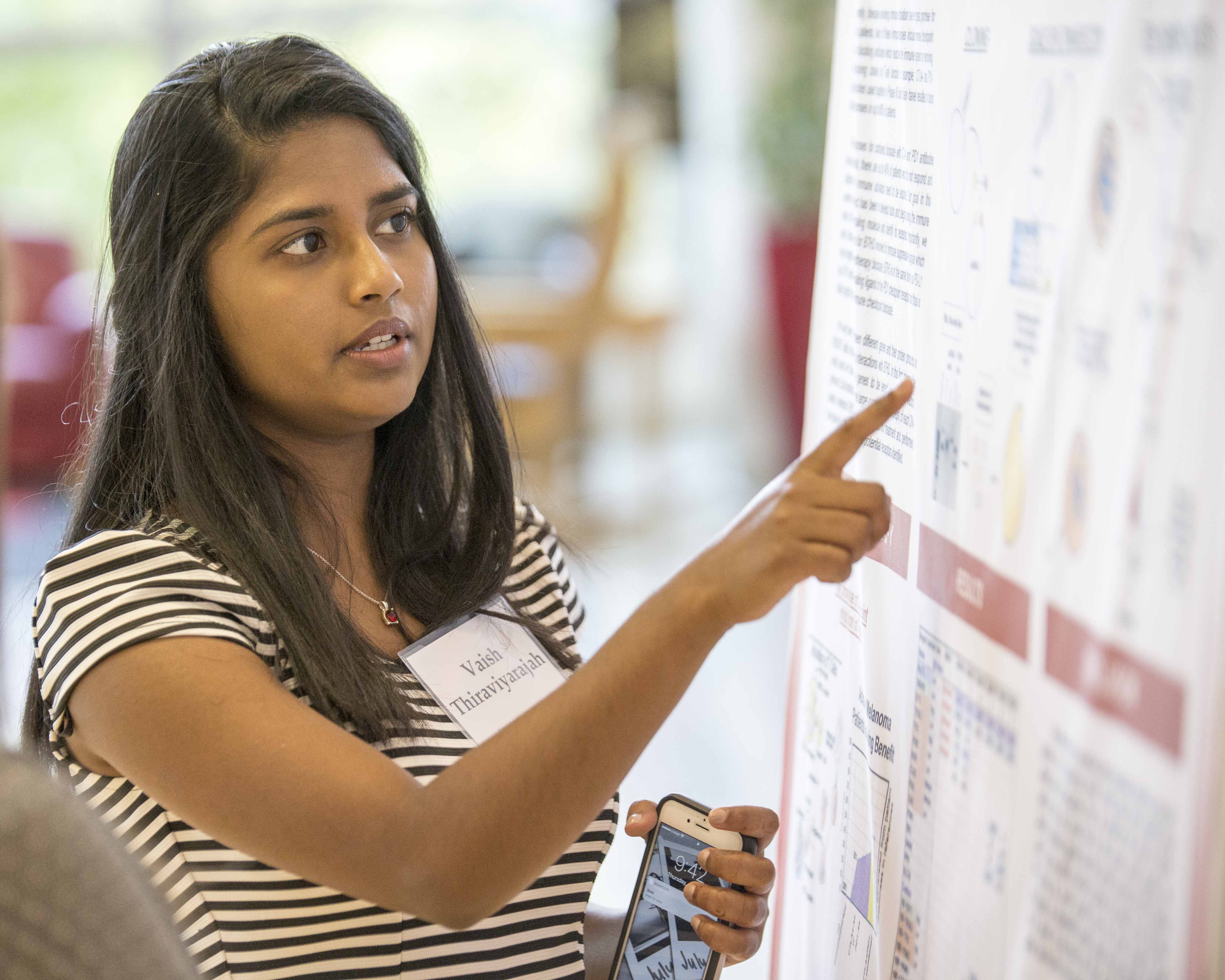 A female student points to a board has she examples her research project. She is wearing a short sleeve blouse with horizontal black stripes.